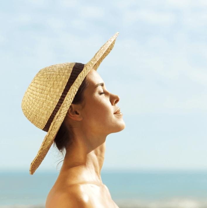 Frau mit Strohhut, die entspannt am Strand steht und das Sonnenlicht genießt.