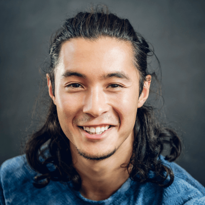 Lachender Mann mit langen Haaren, der in die Kamera lächelt, trägt ein blaues Shirt.