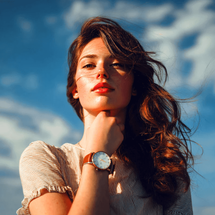 Frau mit rotem Lippenstift und Armbanduhr vor einem wolkigen Himmel