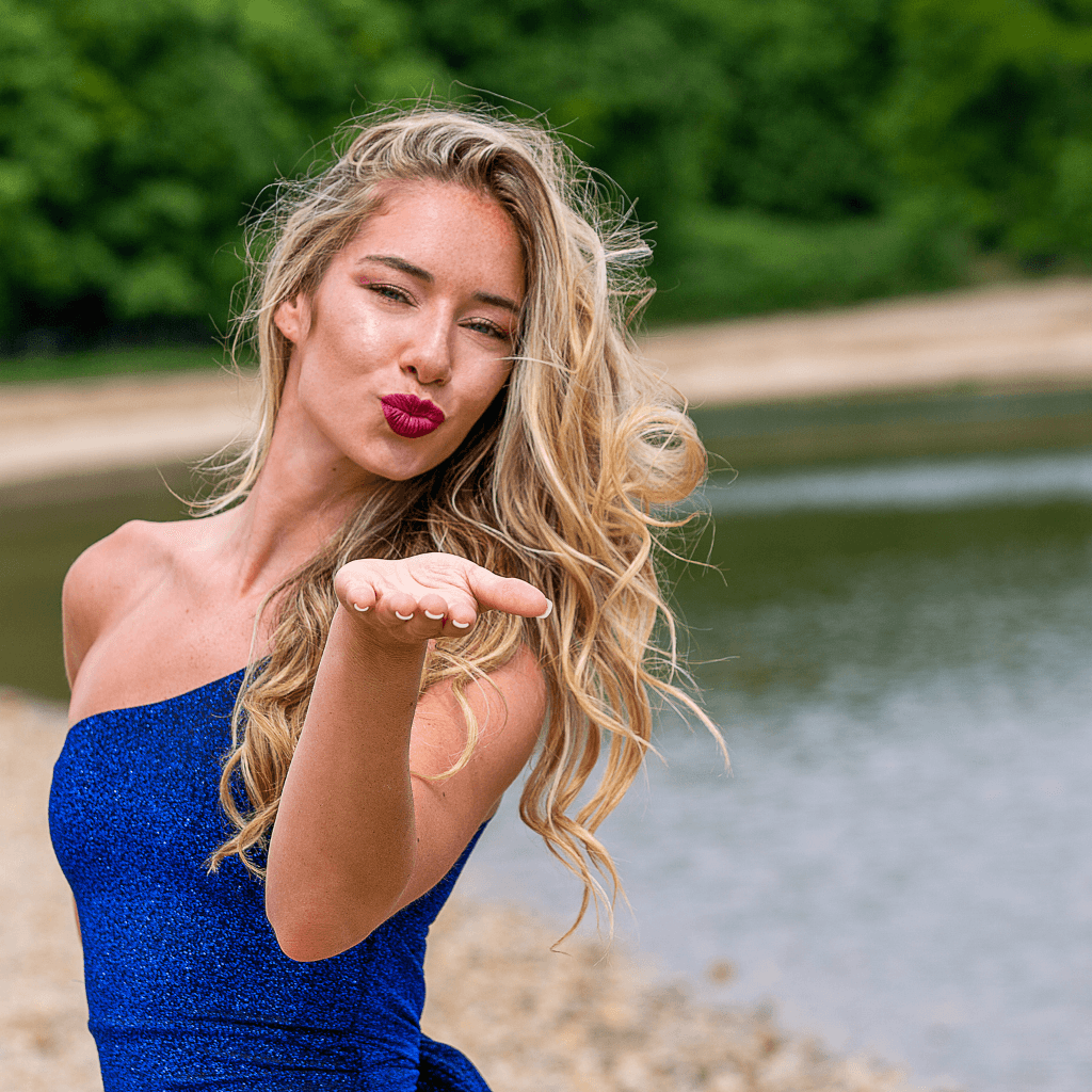 Frau in blauer Kleidung, die einen Kuss wirft, vor einem ruhigen Gewässer. Ideal für Beauty- und Mode-Kampagnen.